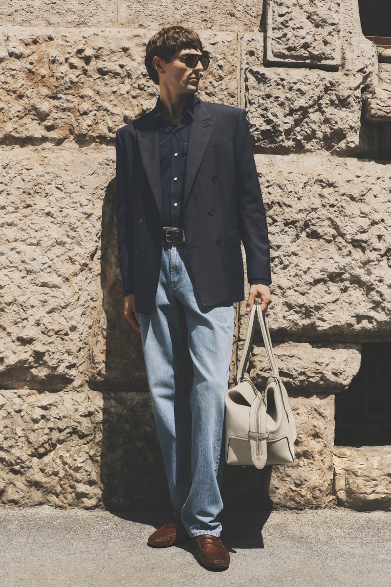Model Joshua (Tomorrow is Another Day) in dark sunglasses wears a navy double-breasted blazer over a navy shirt, paired with light wash blue jeans, brown tassel loafers, and a cream leather top-handle bag