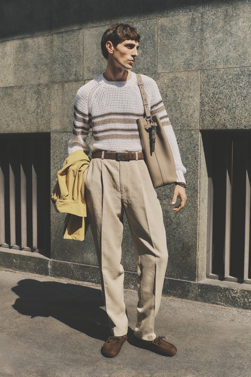 Model Joshua (Tomorrow is Another Day) in a white and tan striped openwork knit sweater, paired with sand pleated trousers, a brown braided belt, brown suede driving loafers, and a taupe leather tote with a yellow jacket draped over the arm