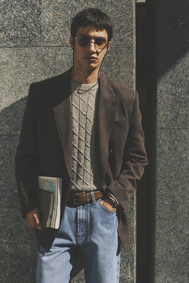 Model Alejandro Albaladejo in tortoise aviator sunglasses wears a chocolate brown blazer over a gray cable-knit sweater, paired with a brown leather belt and light wash blue jeans, holding a folded magazine