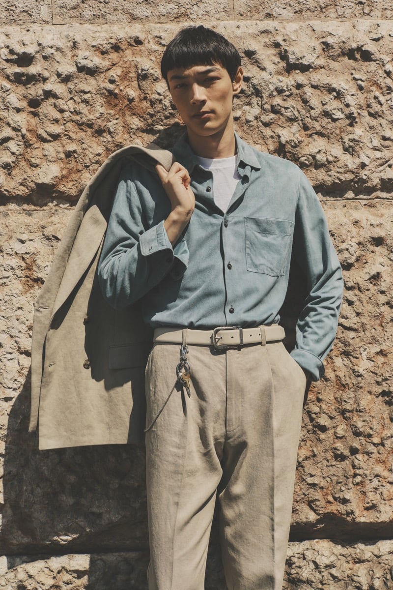 Model Pascal Thulin in a sage blue shirt over a white tee, paired with stone pleated trousers, a leather belt with key chain, and a sand linen blazer carried over the shoulder