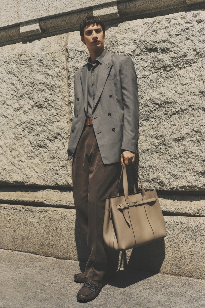 Model Alejandro Albaladejo in a gray Prince of Wales check double-breasted blazer over a shirt, paired with chocolate brown pleated trousers, brown suede loafers, and a large taupe leather tote