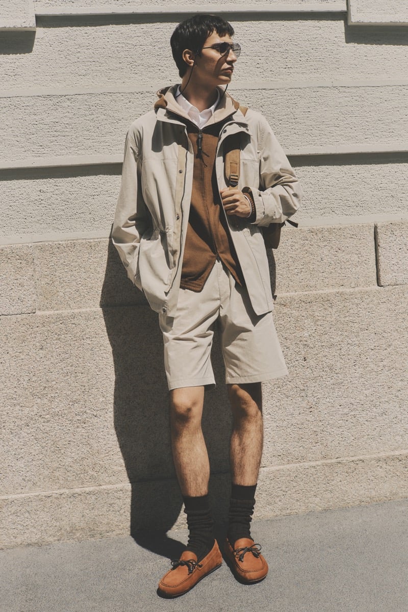 Model Alejandro Albaladejo in slim metal sunglasses wears a stone technical parka over a tan zip jacket and white shirt, paired with matching stone shorts, dark brown socks, and rust suede driving loafers