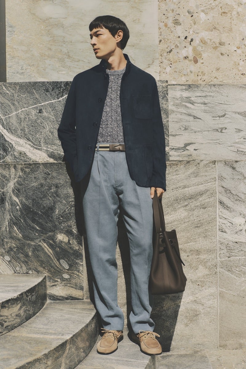 Model Pascal Thulin in a navy chore jacket over a gray marled knit, paired with chambray blue pleated trousers, a brown belt, tan suede boat shoes, and a taupe leather tote