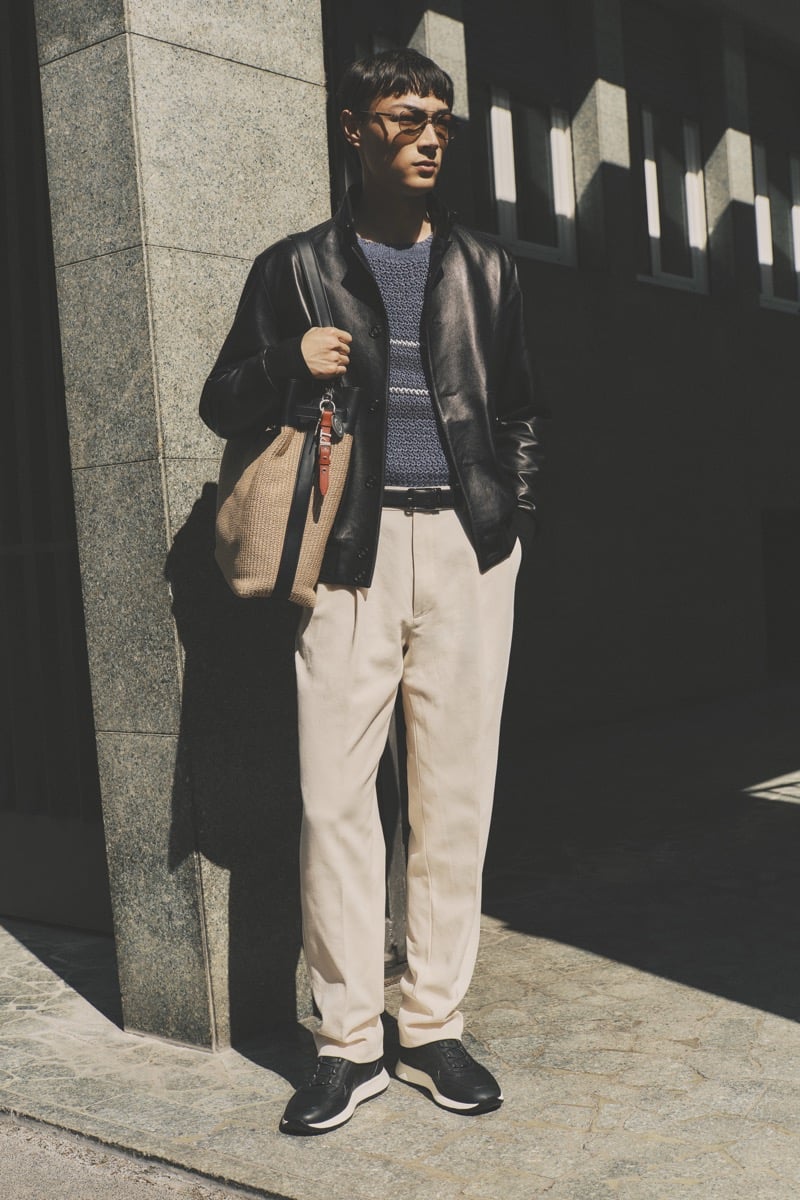 Model Pascal Thulin in tortoise glasses wears a black leather shirt jacket over a blue marled knit, paired with cream pleated trousers, black leather sneakers, and a woven sand tote with leather trim