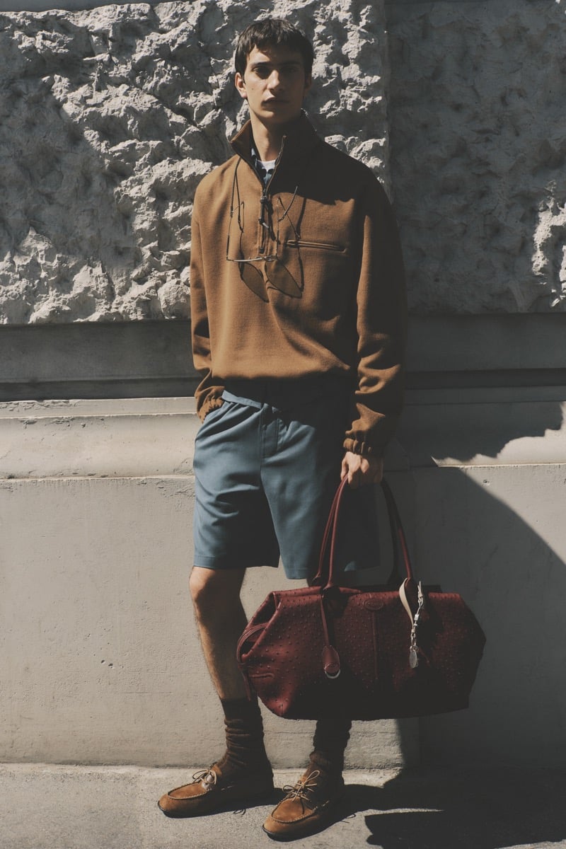 Model Alejandro Albaladejo in a tobacco brown wool quarter-zip pullover with metal sunglasses tucked at the chest, paired with slate blue shorts, brown ribbed socks, tan suede moccasins, and a burgundy ostrich-embossed leather holdall