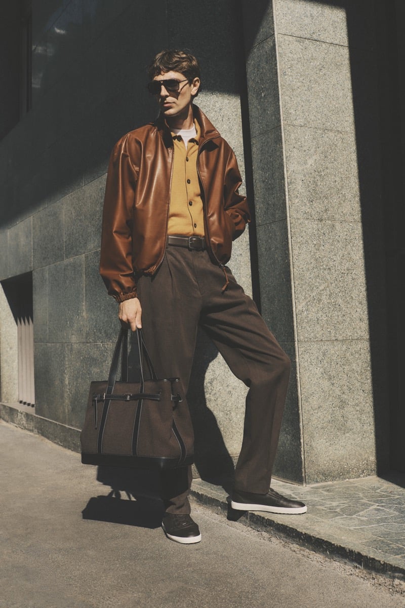 Model Joshua (Tomorrow is Another Day) in dark aviator sunglasses wears a cognac leather bomber jacket over a mustard yellow polo, paired with chocolate brown pleated trousers, dark brown sneakers, and a brown leather and canvas tote