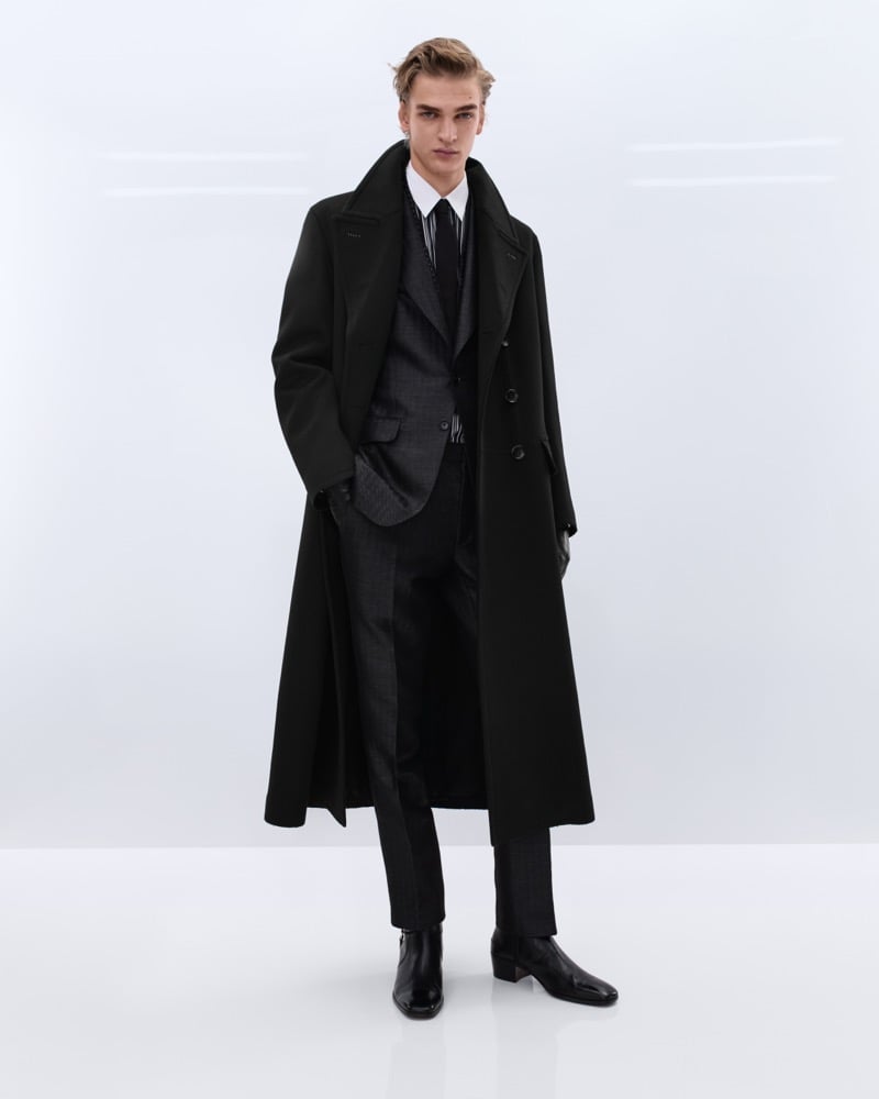 Dario Tonin wears a long black double-breasted wool overcoat over a charcoal Prince of Wales check suit with a white shirt and dark striped tie, holding black leather gloves