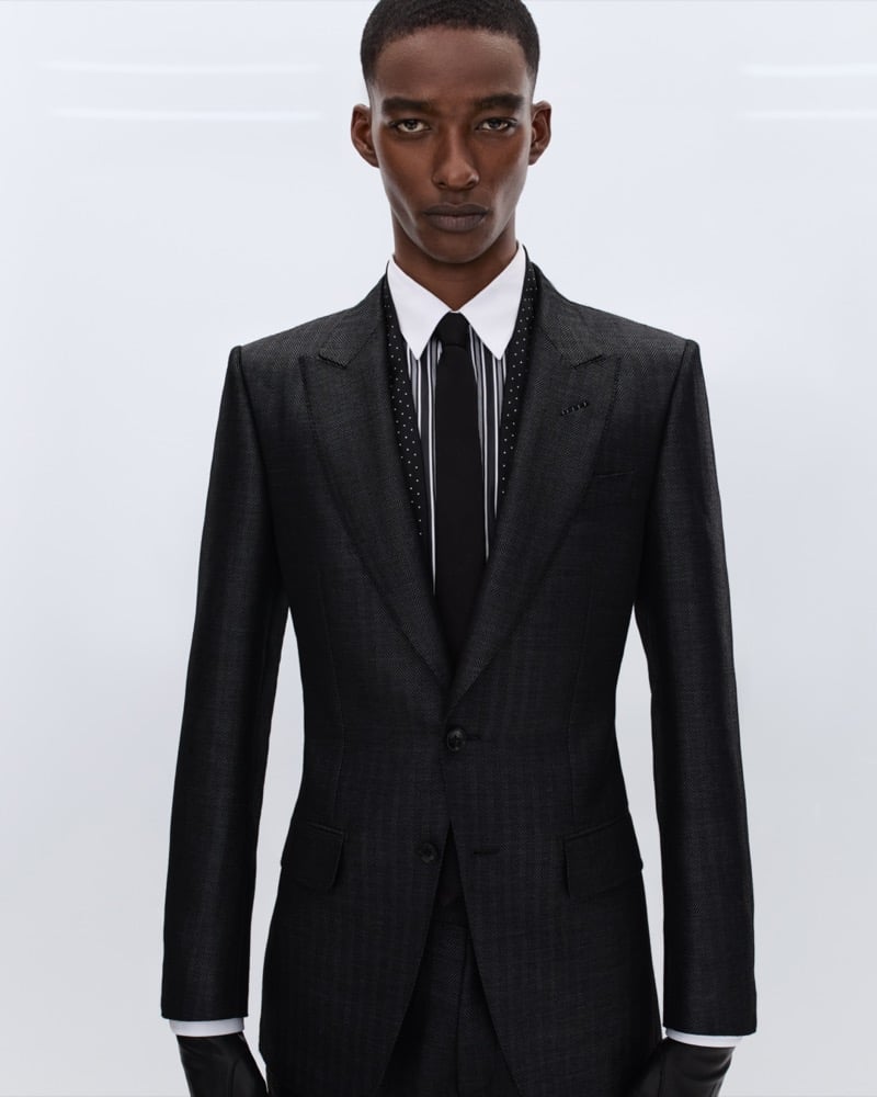 Samuel Elie wears a charcoal single-breasted suit over a white shirt with a black satin tie and a black polka dot and striped scarf collar