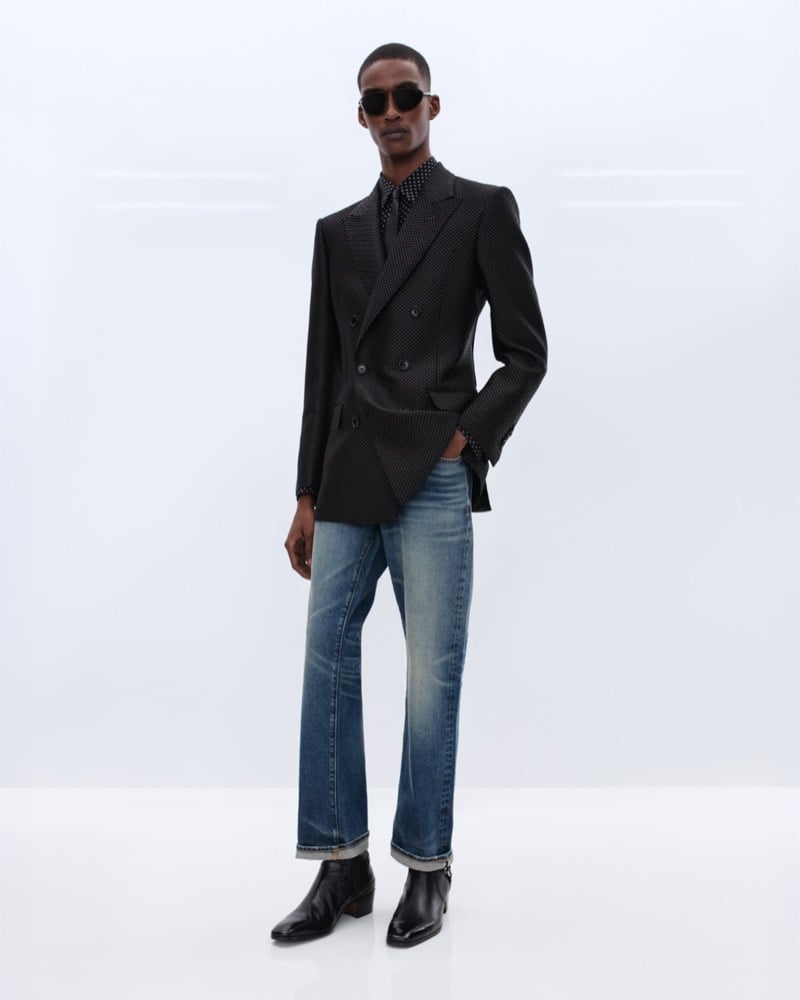 Samuel Elie wears black sunglasses, a black double-breasted micro-dot blazer over a black polka dot shirt with washed indigo jeans and black leather Chelsea boots