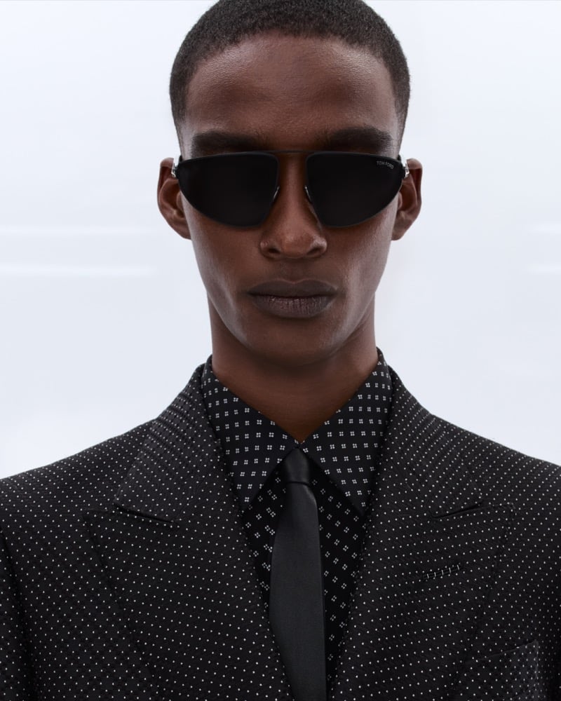 Samuel Elie wears black aviator sunglasses with a black polka dot shirt, black tie, and a black micro-dot suit jacket