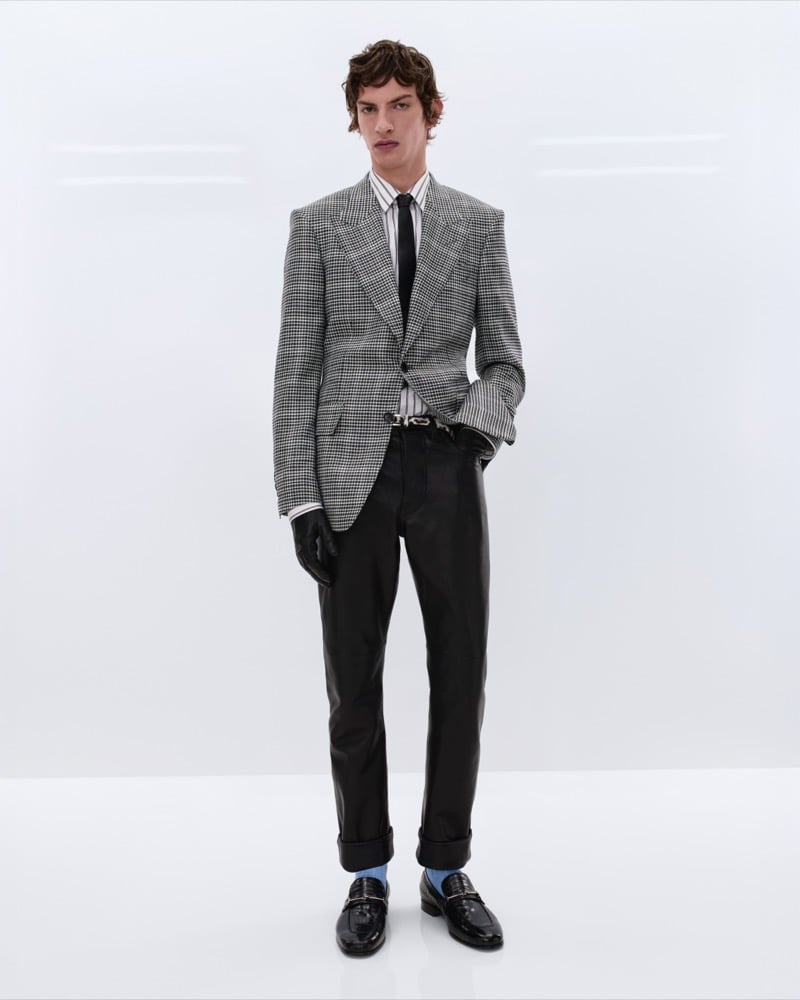 Elliot Belot wears a black and white houndstooth blazer over a striped shirt with a black tie, black leather trousers, and black leather penny loafers with blue socks