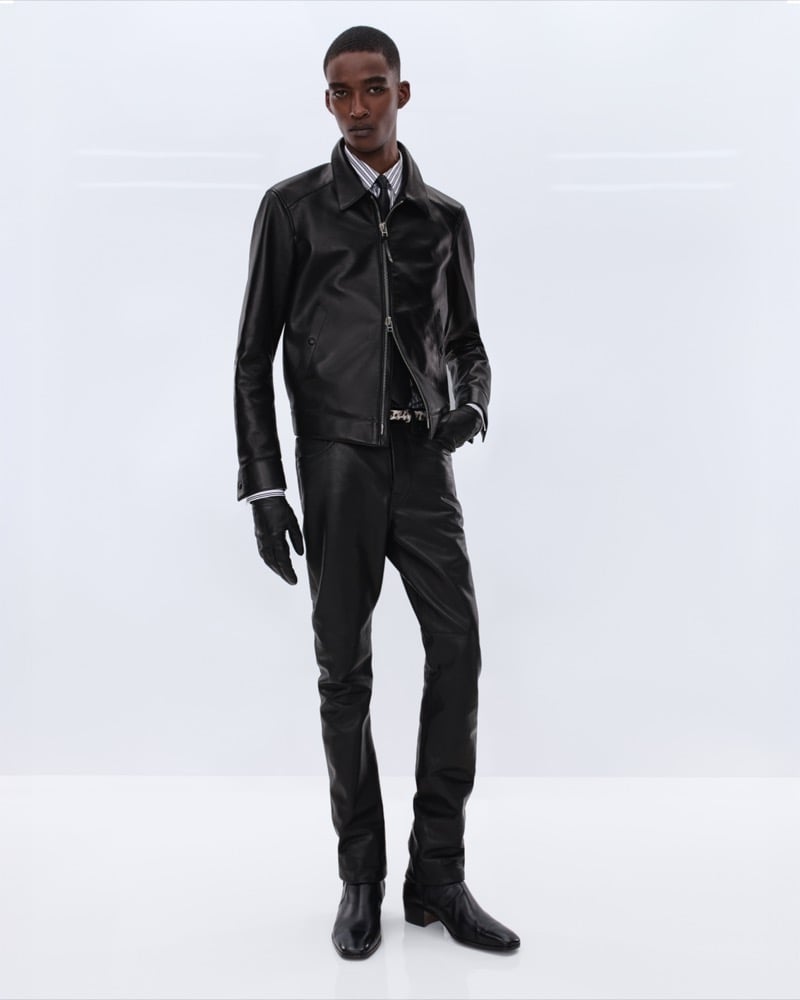 Samuel Elie wears a black leather zip-front jacket over a striped shirt with black leather trousers and black leather gloves and Chelsea boots