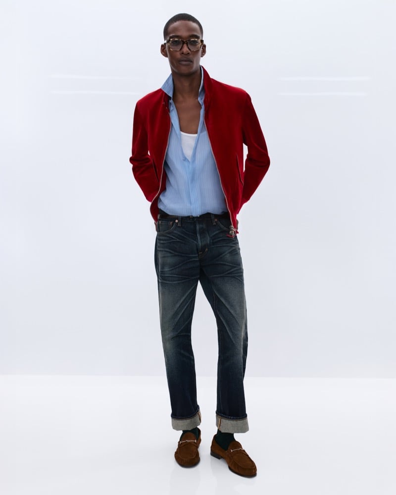 Samuel Elie wears tortoiseshell glasses and a crimson velvet harrington jacket over an unbuttoned blue striped shirt with a white tank and faded indigo jeans with brown suede horsebit loafers