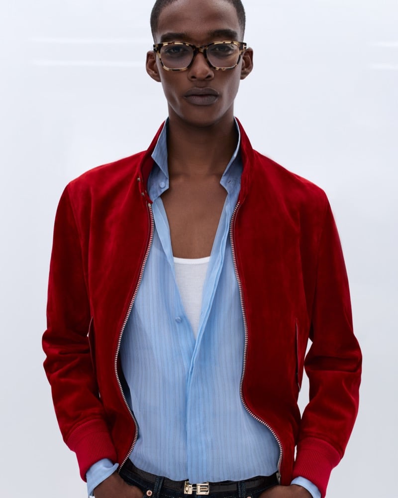Samuel Elie wears tortoiseshell glasses and an unzipped crimson suede bomber jacket over an open pale blue shirt and white ribbed tank with a gold logo belt