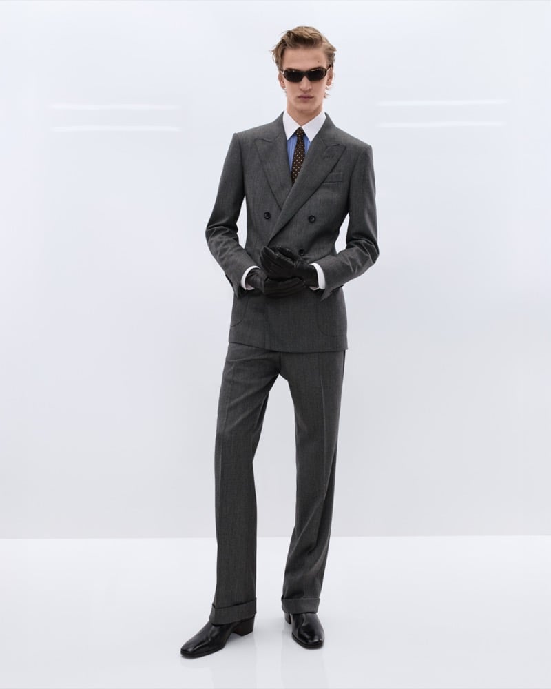 Dario Tonin wears black sunglasses, a grey double-breasted wool suit over a blue shirt with a brown polka dot tie, and black leather driving gloves