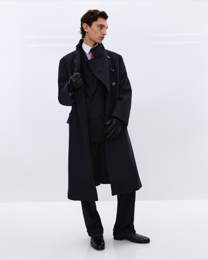 Cirillo wears a long navy double-breasted wool overcoat over a charcoal pinstripe suit with a white shirt and red tie, holding black leather gloves