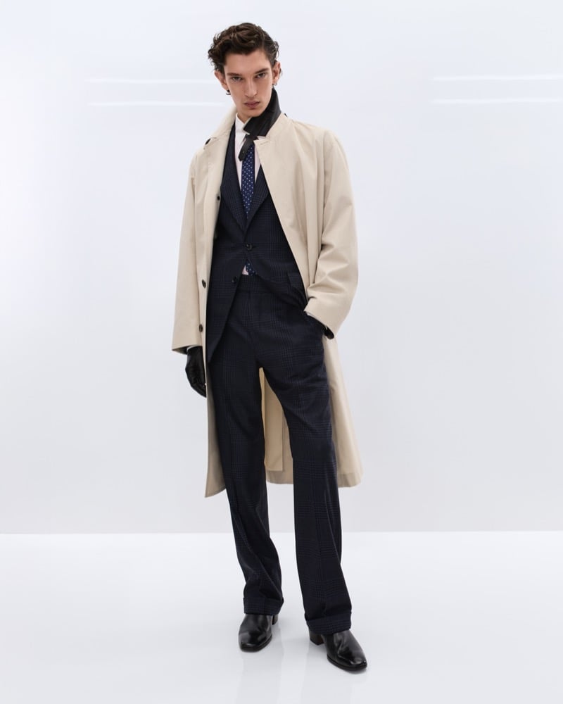 Cirillo wears a beige cotton trench coat over a navy Prince of Wales check three-piece suit with a pink shirt and navy polka dot tie, holding black leather gloves