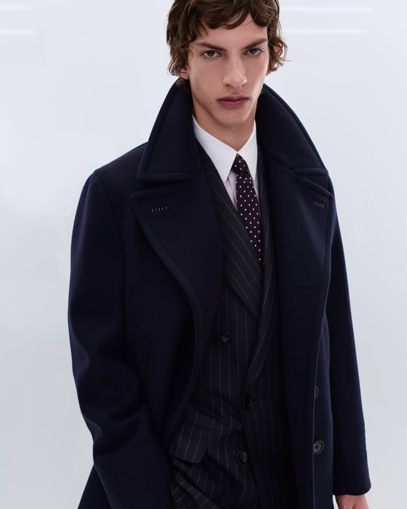 Elliot Belot wears an oversized navy wool peacoat with a wide lapel over a charcoal pinstripe suit, white shirt, and burgundy polka dot tie