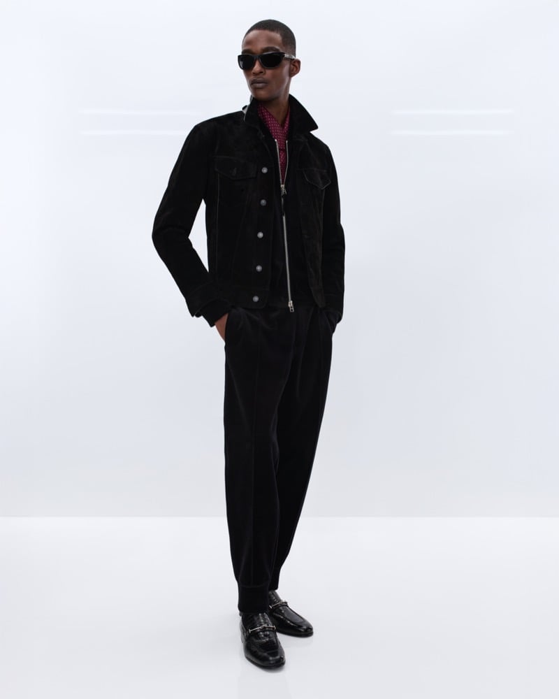 Samuel Elie wears black sunglasses and a black suede trucker jacket over a burgundy polka dot shirt with black corduroy pleated trousers and black leather horsebit loafers
