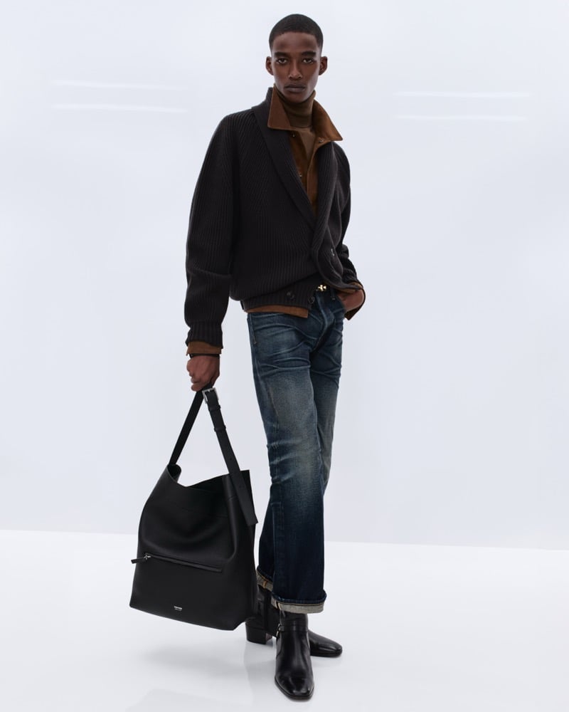 Samuel Elie wears a black ribbed wool cardigan over a brown corduroy shirt and rust turtleneck with washed indigo bootcut jeans, holding a large black leather Tom Ford tote