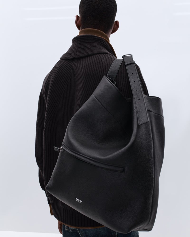 Samuel Elie wears a black ribbed knit sweater over brown corduroy shirting, slung with a large black pebbled leather Tom Ford tote bag