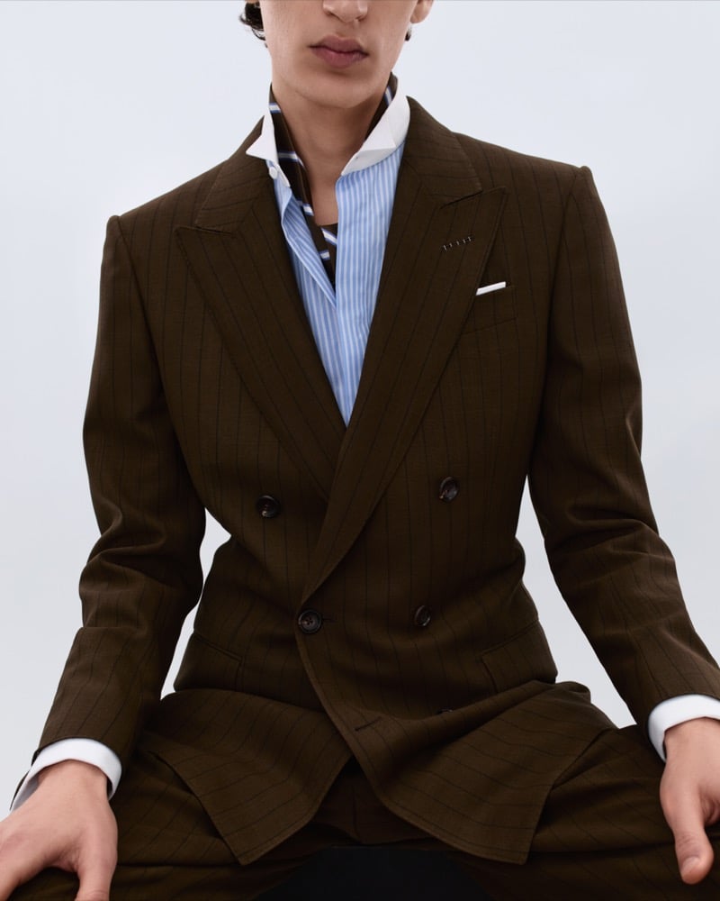 Cirillo wears a brown chalk stripe double-breasted suit jacket over a blue striped shirt and a black and white striped scarf at the open collar with a white pocket square