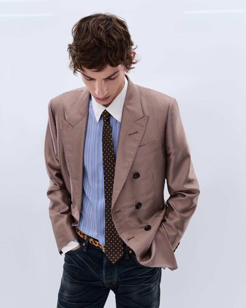 Elliot Belot wears a camel jacquard double-breasted blazer over a blue striped shirt with a brown polka dot tie and a leopard print belt with washed indigo jeans