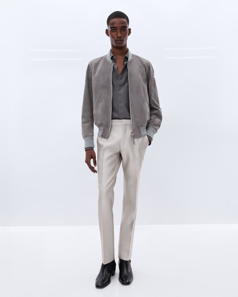 Samuel Elie wears a faded grey suede bomber jacket over a grey silk shirt with cream wool trousers and black leather Chelsea boots