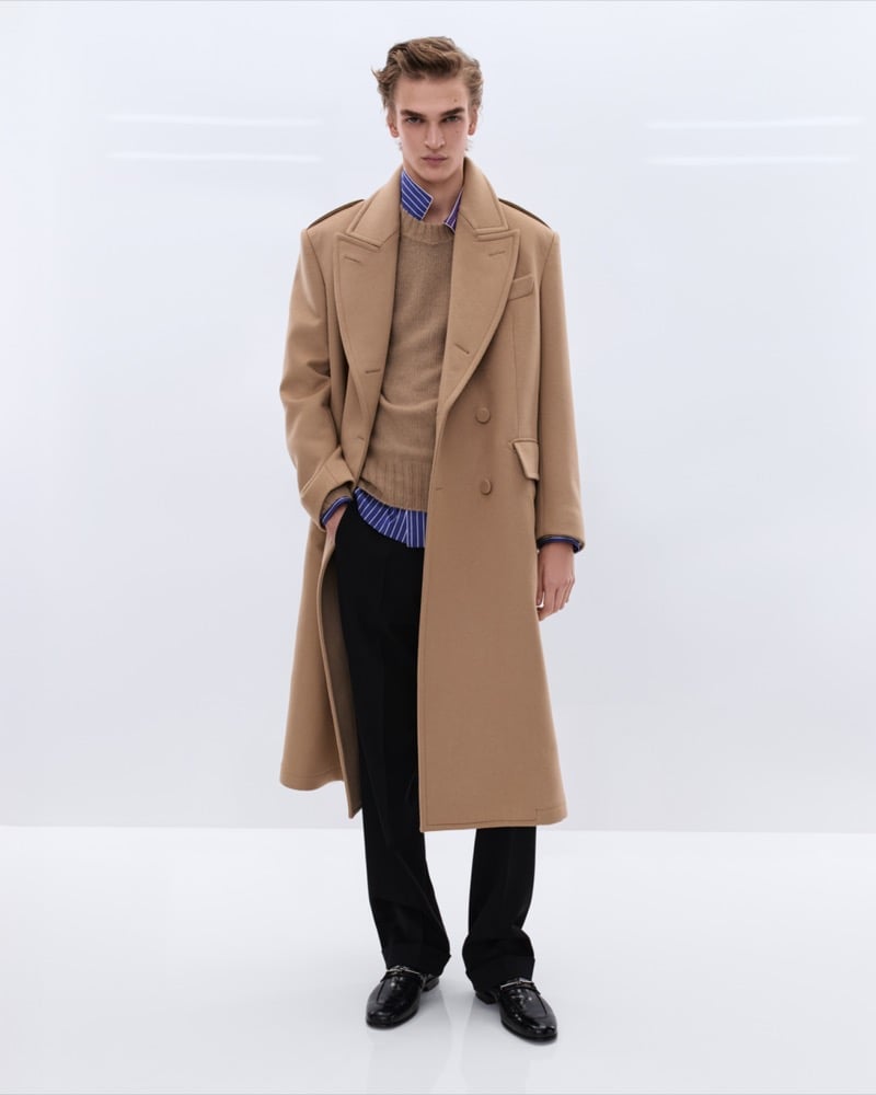 Dario Tonin wears a long camel double-breasted wool overcoat over a tan cashmere sweater and blue striped shirt with black trousers and black leather horsebit loafers