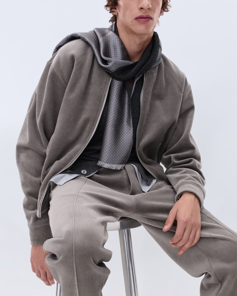 Elliot Belot wears a faded taupe suede bomber jacket with a grey polka dot scarf over a grey waistcoat and matching taupe trousers, seated on a stool