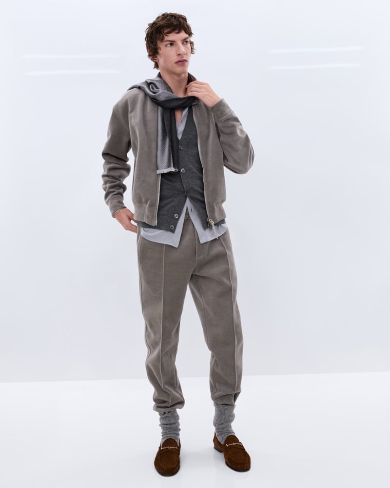 Elliot Belot wears a faded taupe suede bomber jacket with a grey polka dot scarf over a grey waistcoat and taupe trousers with grey wool socks and brown suede horsebit loafers