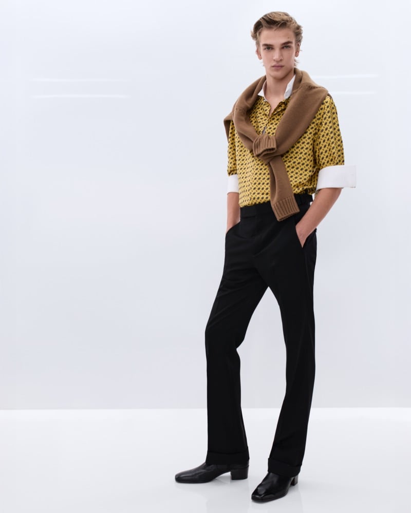 Dario Tonin wears a yellow patterned short-sleeve silk shirt over a white tank with a tan cashmere sweater knotted at the shoulders and black trousers with black leather Chelsea boots