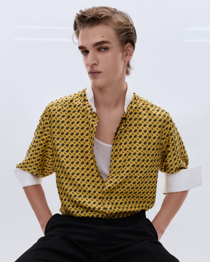Dario Tonin wears a yellow paisley silk camp shirt with rolled cuffs over a white ribbed tank, hands in pockets