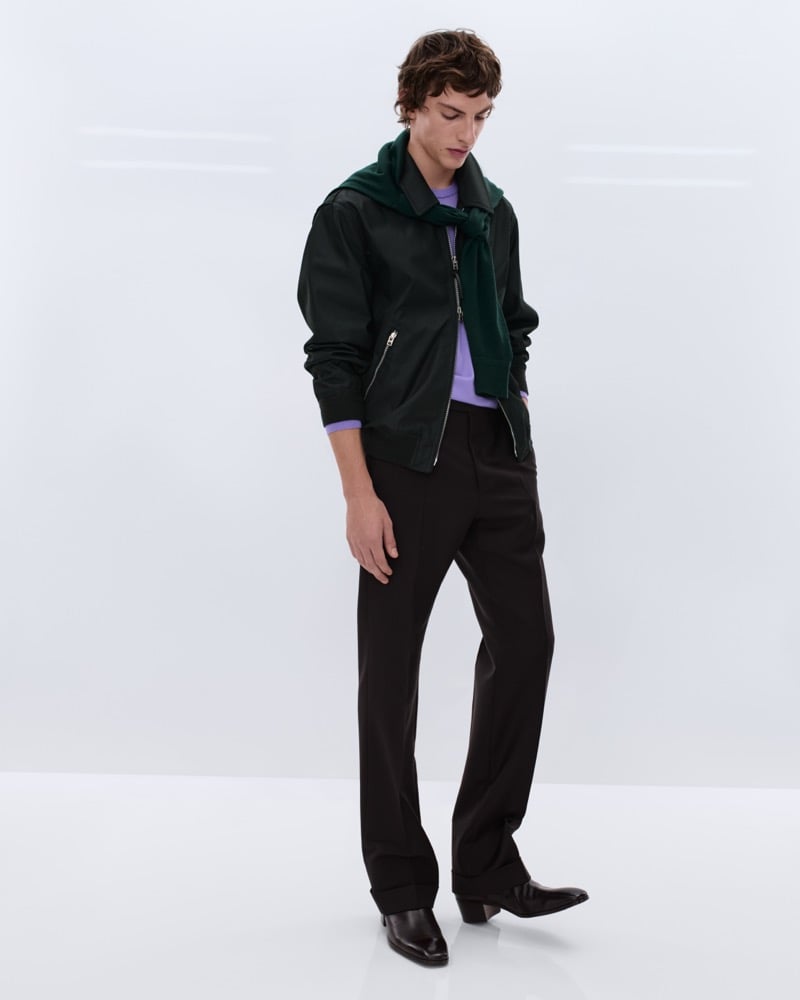 Elliot Belot wears a dark green satin bomber jacket over a lavender shirt with a forest green hooded sweatshirt tied at the shoulders and black wool trousers with black leather Chelsea boots