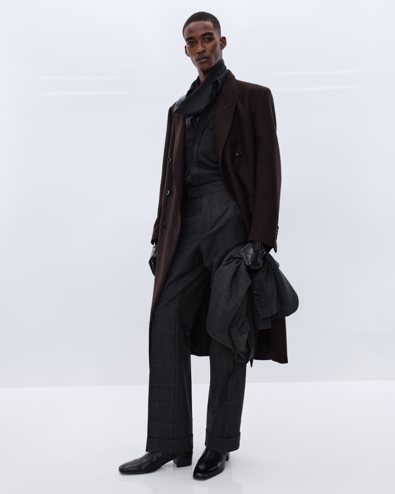 Samuel Elie wears a brown wool overcoat over a black silk shirt and a brown polka dot scarf with grey Prince of Wales check trousers, holding a charcoal jacket and black leather gloves