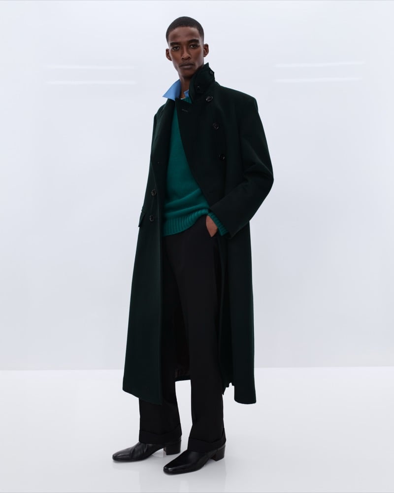 Elliot Belot wears a long forest green wool double-breasted overcoat over a teal cashmere crewneck sweater and blue shirt with black trousers and black leather Chelsea boots