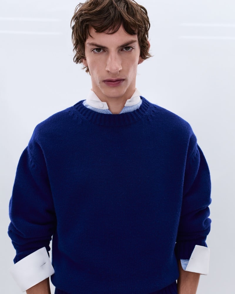 Elliot Belot wears a cobalt blue wool crewneck sweater over a white shirt with oversized cuffs and a blue striped collar