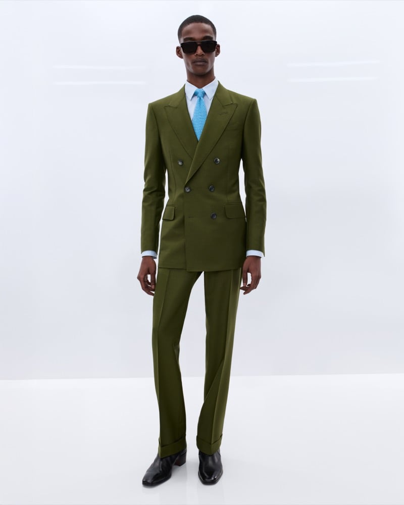 Samuel Elie wears black sunglasses and an olive green double-breasted suit over a pale blue shirt with a textured sky blue tie and black leather Chelsea boots