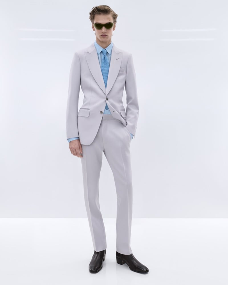 Dario Tonin wears amber-tinted aviator sunglasses with a pale grey single-breasted suit over a pale blue shirt and textured sky blue tie with brown leather Chelsea boots