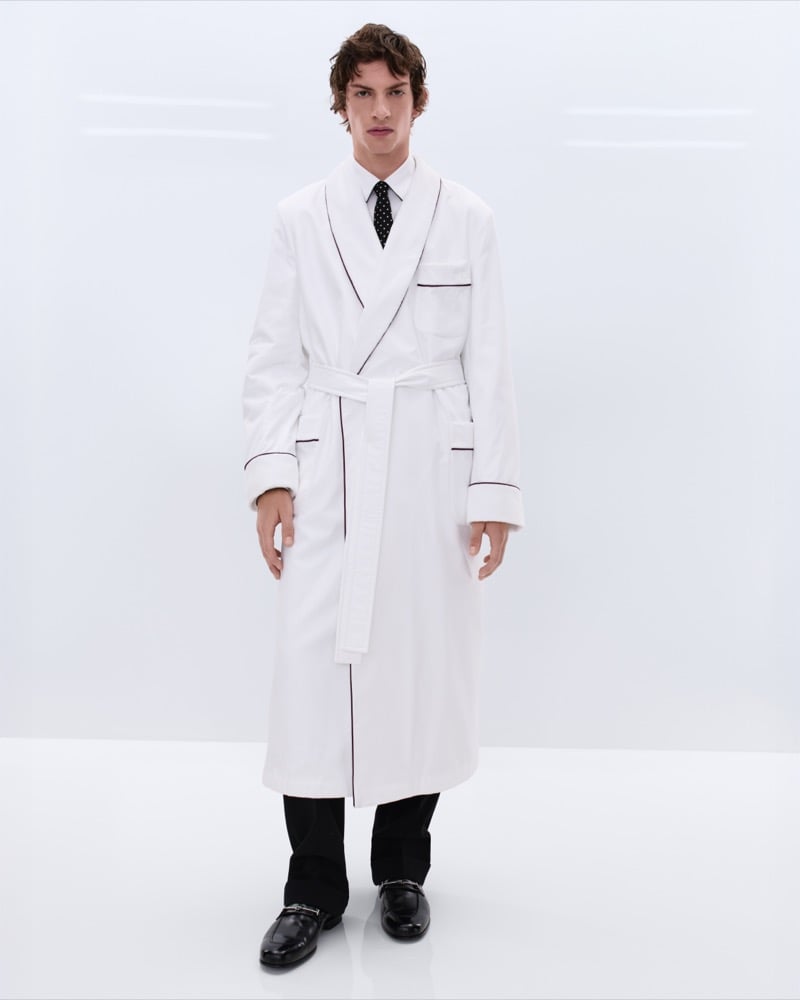 Elliot Belot wears a white cotton dressing gown with burgundy piping over a white shirt and black polka dot tie with black trousers and black leather horsebit loafers