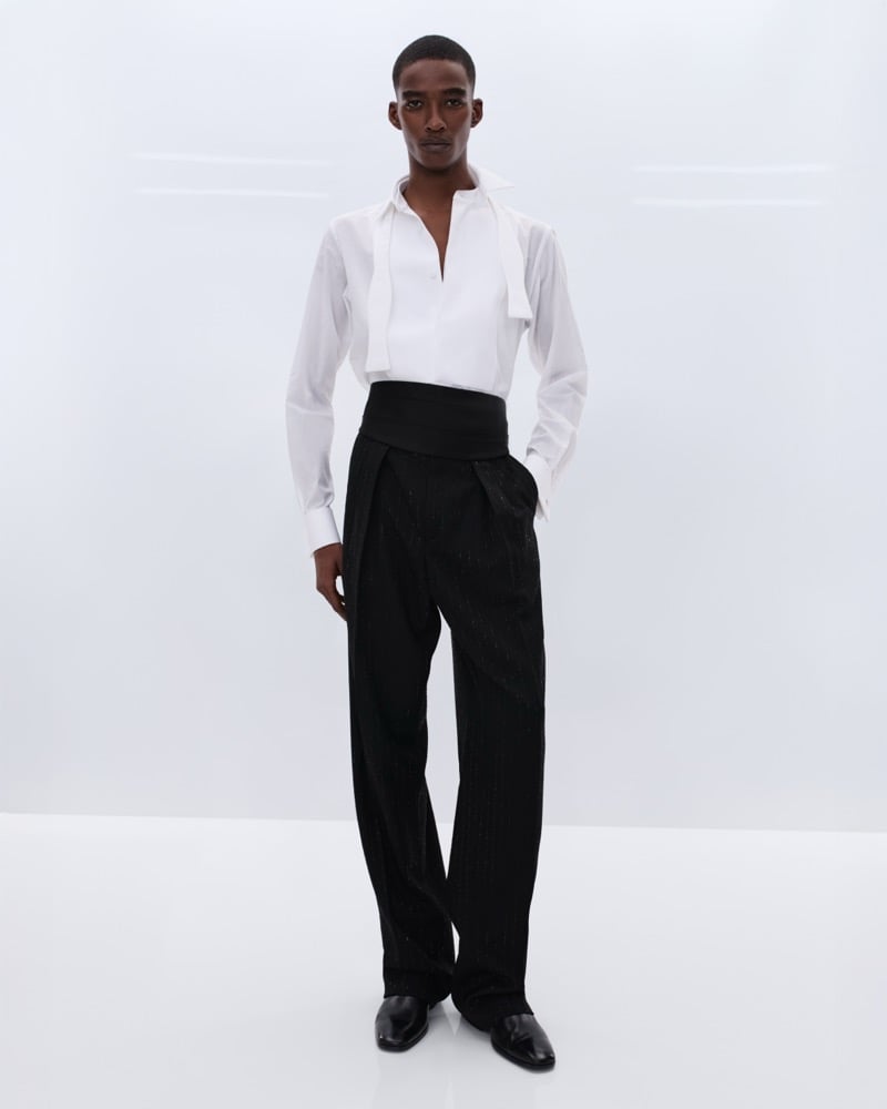 Samuel Elie wears a white silk shirt with a soft neck tie sash and high-waisted black sequin pinstripe trousers with a wide black cummerbund and black leather Chelsea boots