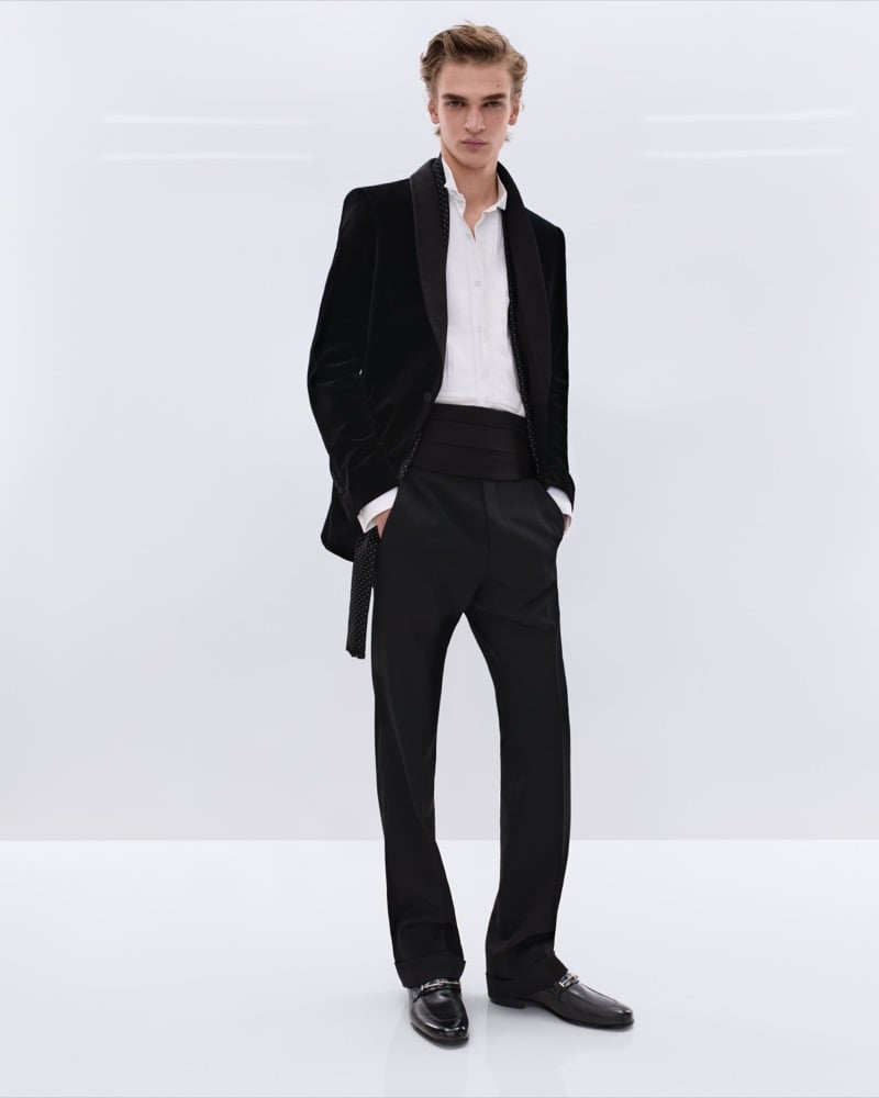 Dario Tonin wears a black velvet shawl collar dinner jacket with beaded trim over a white shirt with high-waisted black trousers, a wide black cummerbund, and black leather horsebit loafers