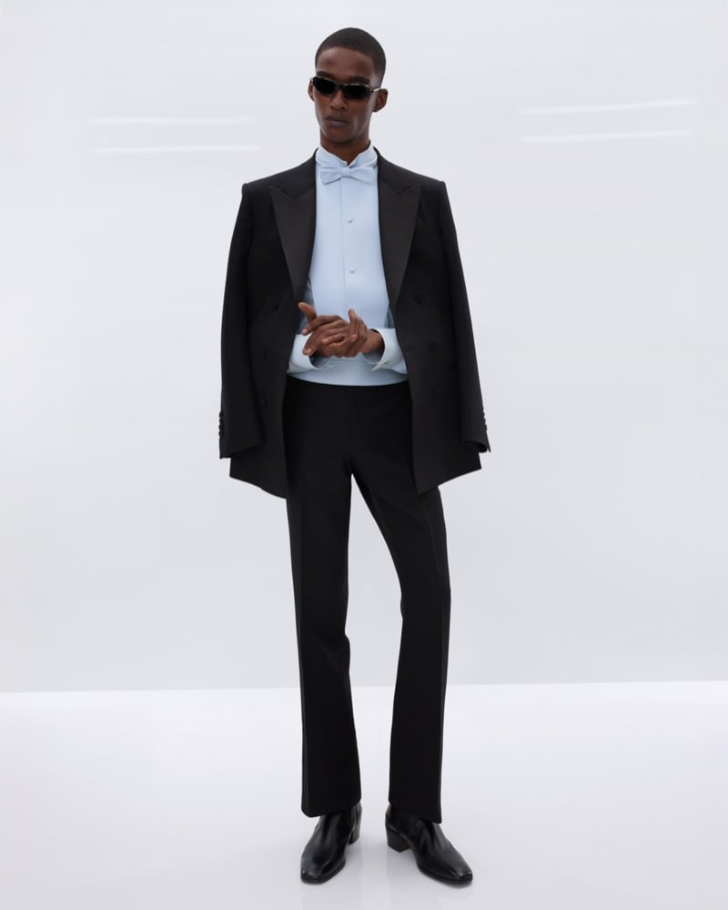 Samuel Elie wears black sunglasses and a black peak lapel tuxedo jacket draped over the shoulders over a pale blue tuxedo shirt with a pale blue bow tie and black trousers with black leather Chelsea boots