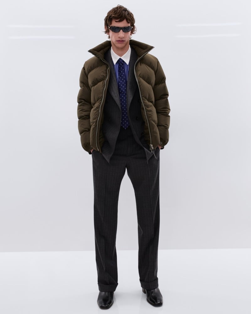 Elliot Belot wears black sunglasses and an olive green down puffer jacket over a charcoal pinstripe double-breasted suit with a white shirt and navy polka dot tie