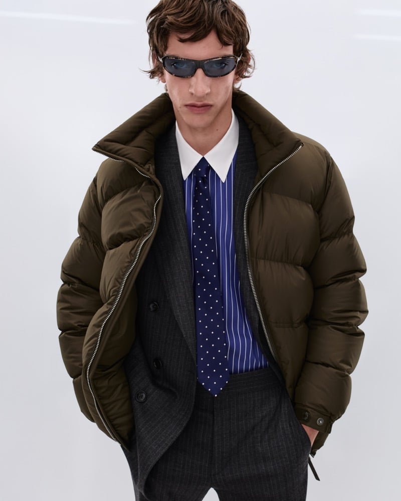 Elliot Belot wears black sunglasses with an olive green quilted down puffer jacket over a charcoal pinstripe suit, blue and white striped shirt, and navy polka dot tie