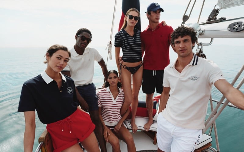Models Hamid Onifade, Kai Paula, and Francisco Henriques pose with female cast members aboard a sailing schooner for Tommy Hilfiger summer 2026, mixing tipped polos, a striped knit polo, swim bottoms, red shorts, and white shorts against the open sea