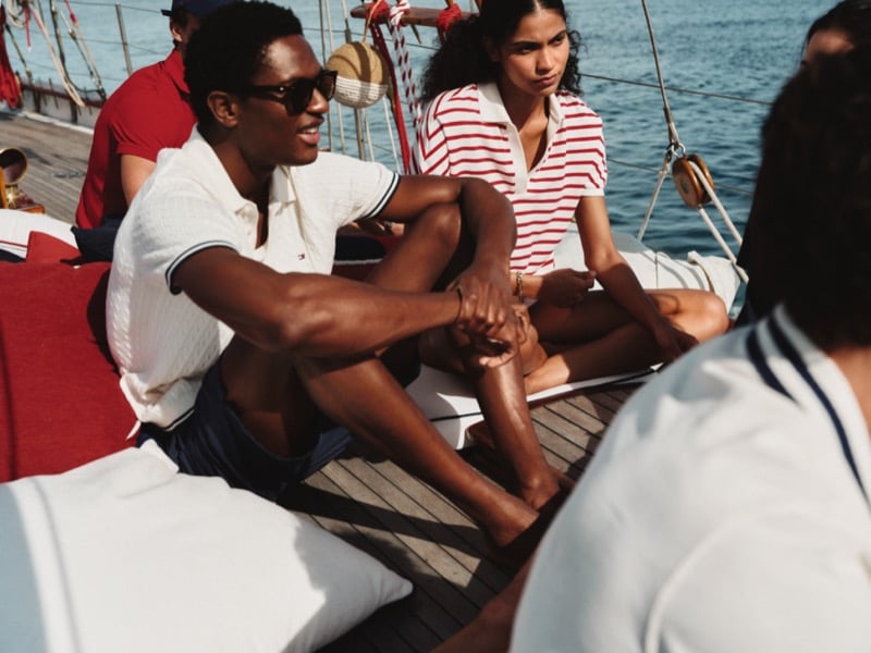 Hamid Onifade and a female cast member share a quiet moment on the schooner deck for Tommy Hilfiger summer 2026, his tipped cable-stitch polo styled with swim shorts beside her red and white striped polo top