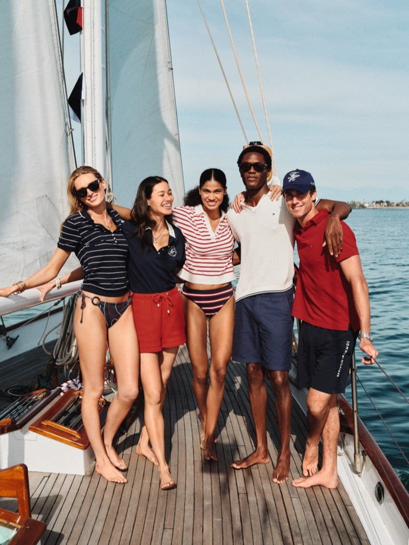 Tommy Hilfiger summer 2026 group portrait on a sailboat deck features Hamid Onifade in a tipped piqué polo and Kai Paula in a red polo with cap, joined by three female cast members in striped knits, red shorts, and printed swim bottoms