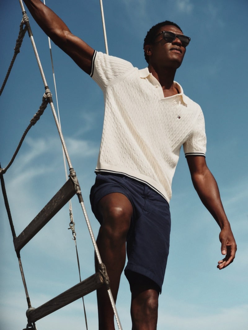 Model Hamid Onifade balances on a rope ladder against a blue sky for Tommy Hilfiger summer 2026, wearing a tipped cable-stitch polo and navy tailored shorts