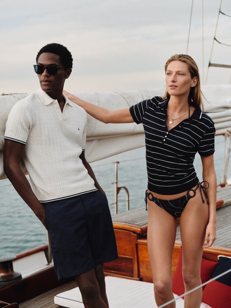 Model Hamid Onifade stands beside a female cast member on the schooner for Tommy Hilfiger summer 2026, his cable-stitch polo paired with navy tailored shorts as she wears a striped polo and printed bikini bottoms